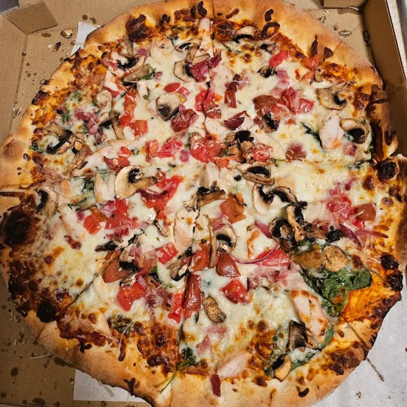 A whole pizza from The Pizza Spot, topped with mushrooms, tomatoes, and cheese in a delivery box.