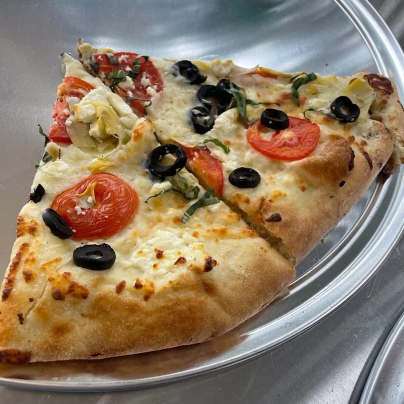 Two slices of The Pizza Spot's white pizza with tomatoes, olives, and basil on a metal tray.
